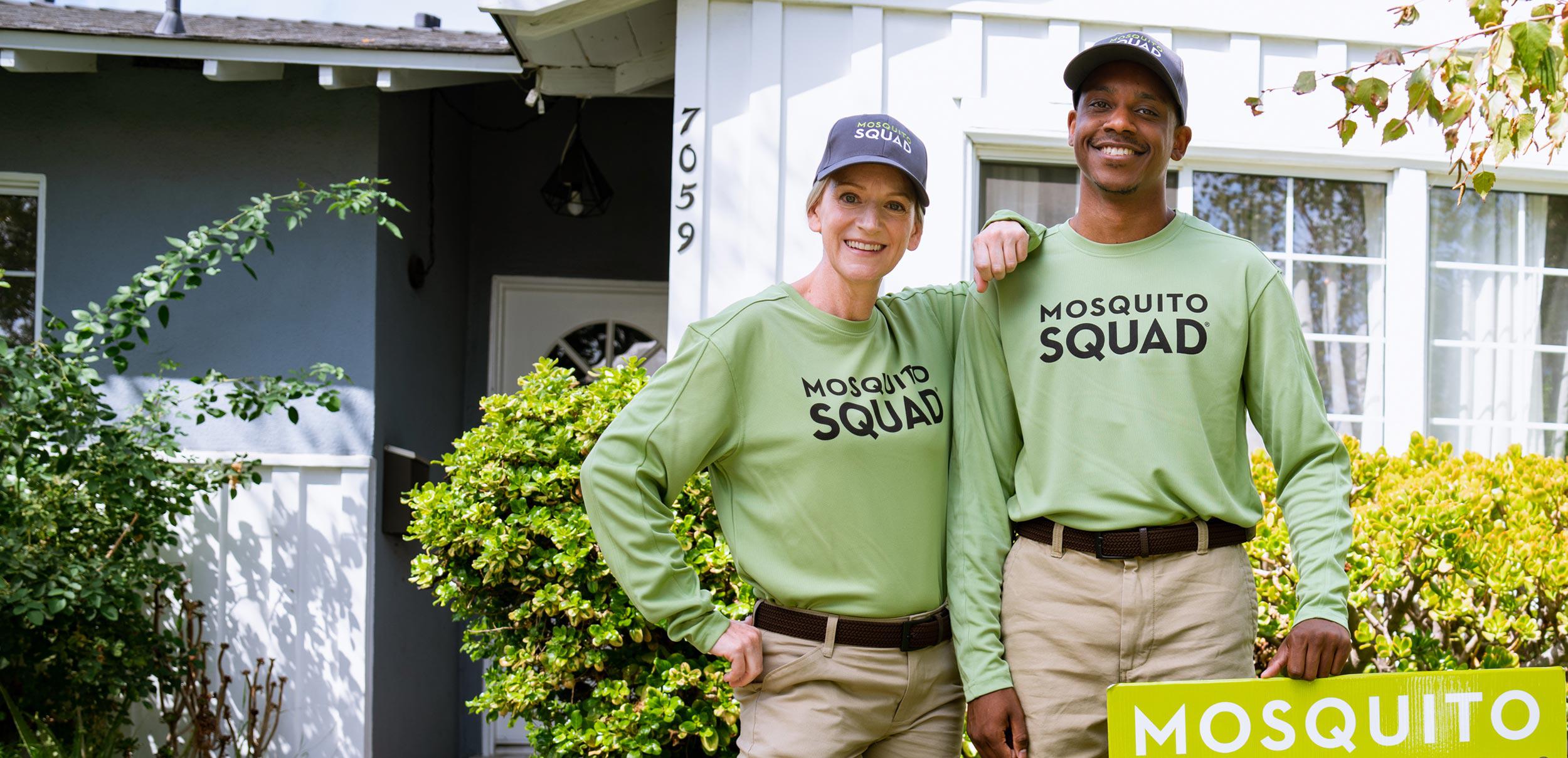 Two Mosquito Squad technicians  in green uniforms and hats, equipped with sprayers, in front of a brick house.