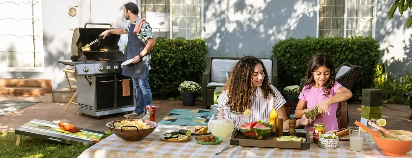 Family enjoying a backyard barbecue: A man is grilling food on a barbecue, while a woman and a young girl prepare the table